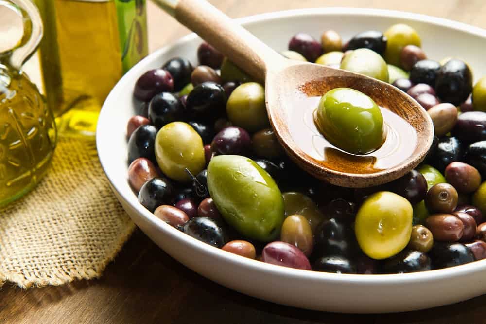 a bowl of green and black olives with one green olive on a spoon