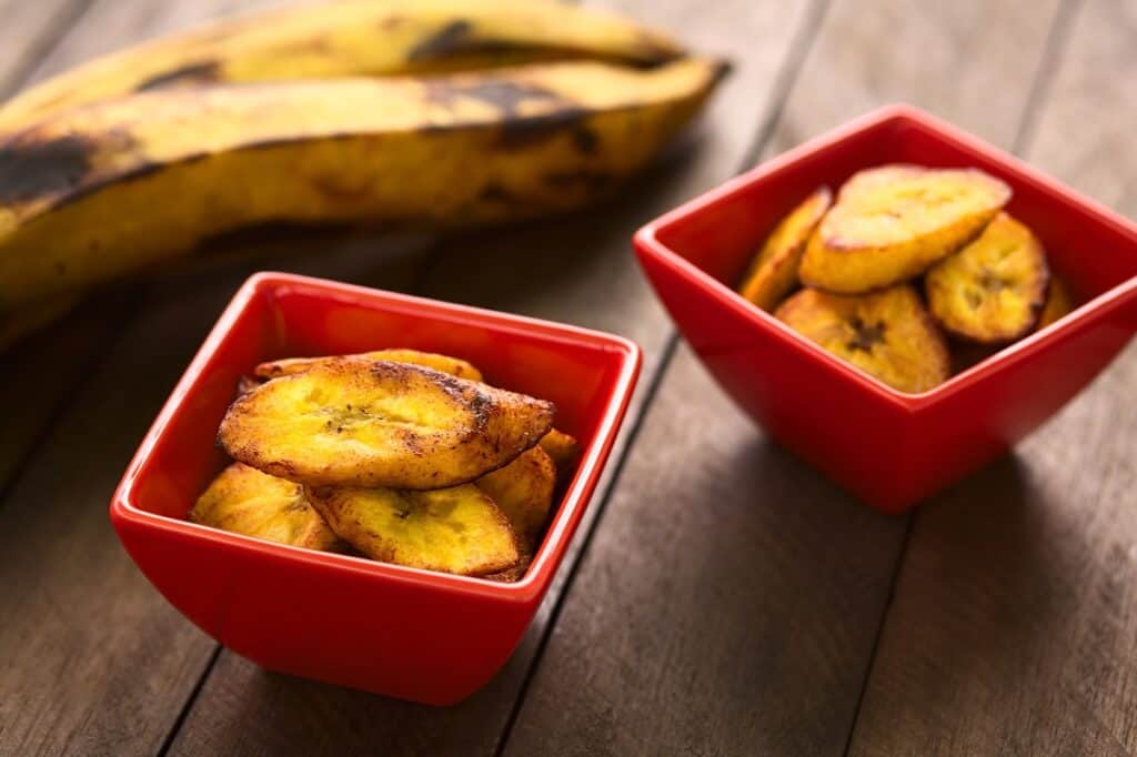 Fried plantain slices in a red bowl, next to a few unpeeled plantains.